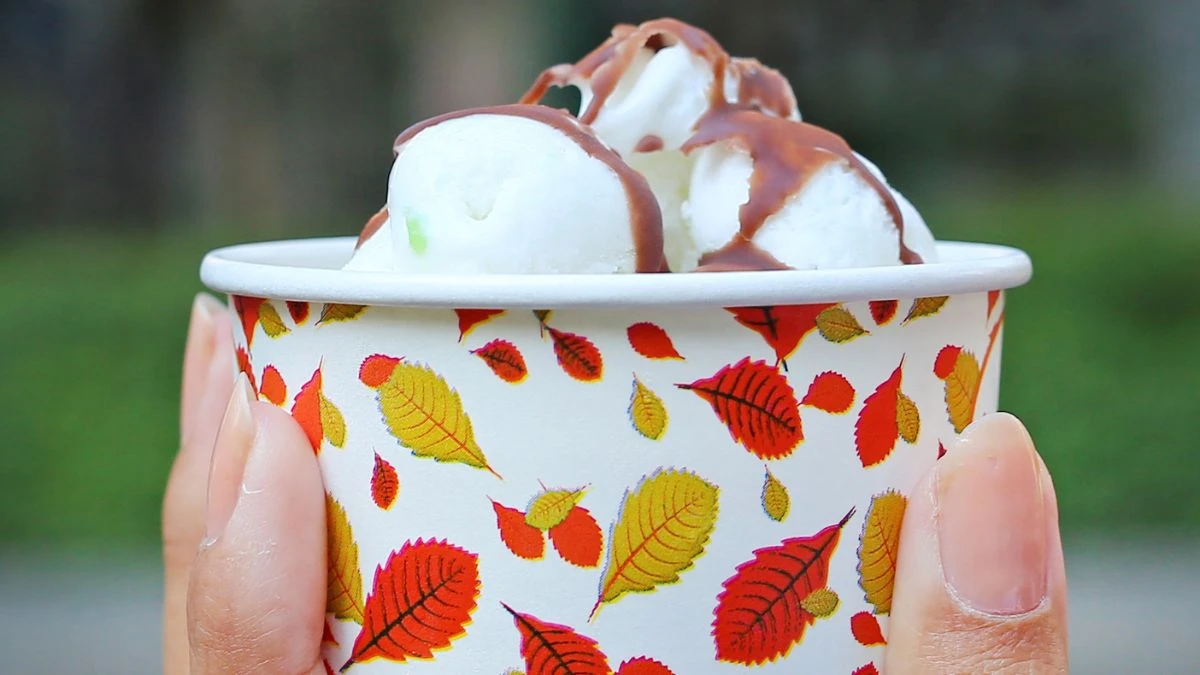 Potes de papel personalizados: como diferenciar sorveteria Uma mão segurando um copo decorado com folhas, cheio de sorvete coberto com calda de chocolate. Uma deliciosa opção de sobremesa para dias quentes.