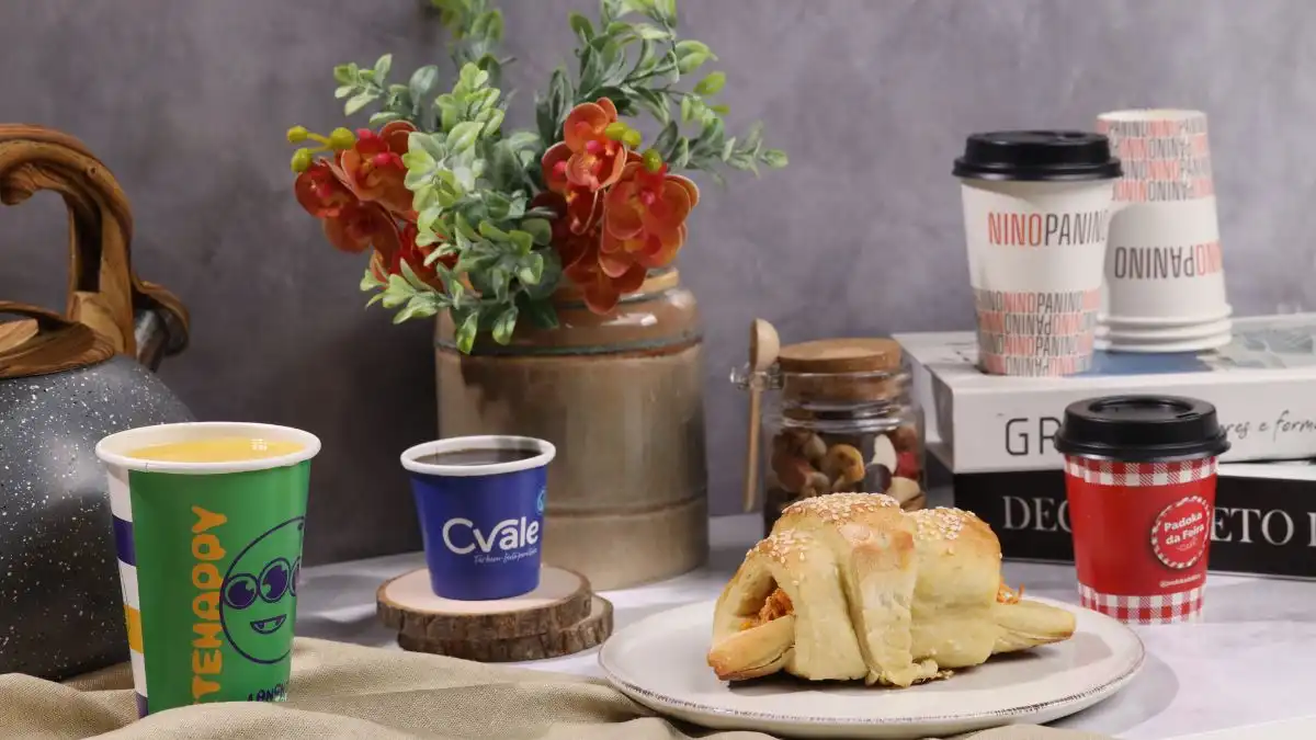 Imagem mostrando uma mesa posta com bebidas, um croissant e uma decoração de flores. A composição destaca produtos e um ambiente aconchegante.