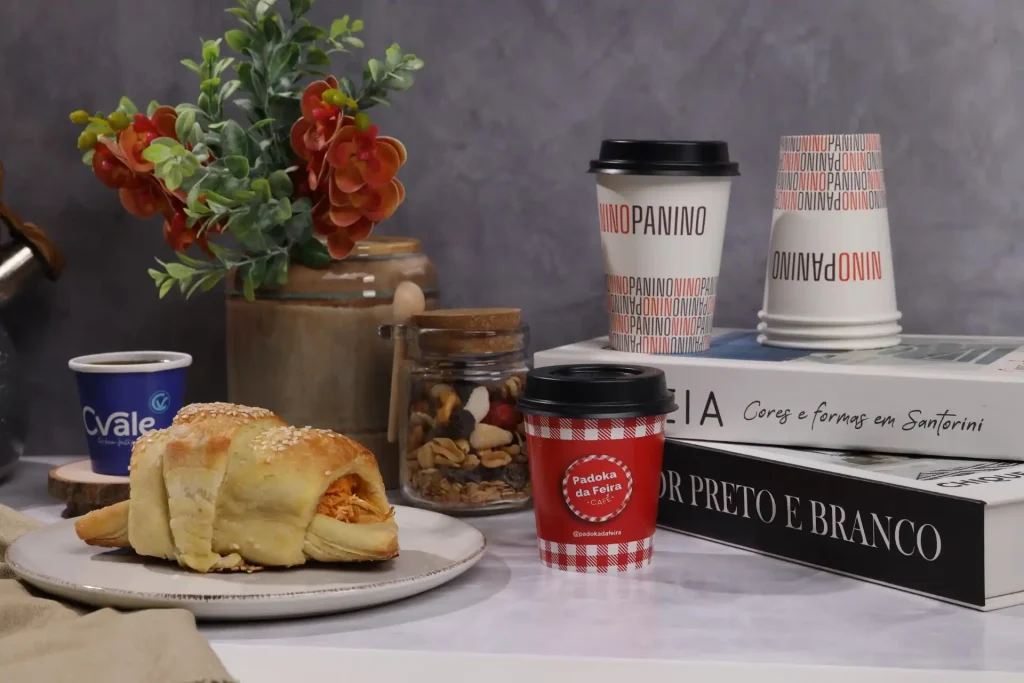Imagem de uma mesa com um croissant recheado, xícaras de café, um vaso de flores, e livros decorativos. Ideal para quem ama café da manhã.