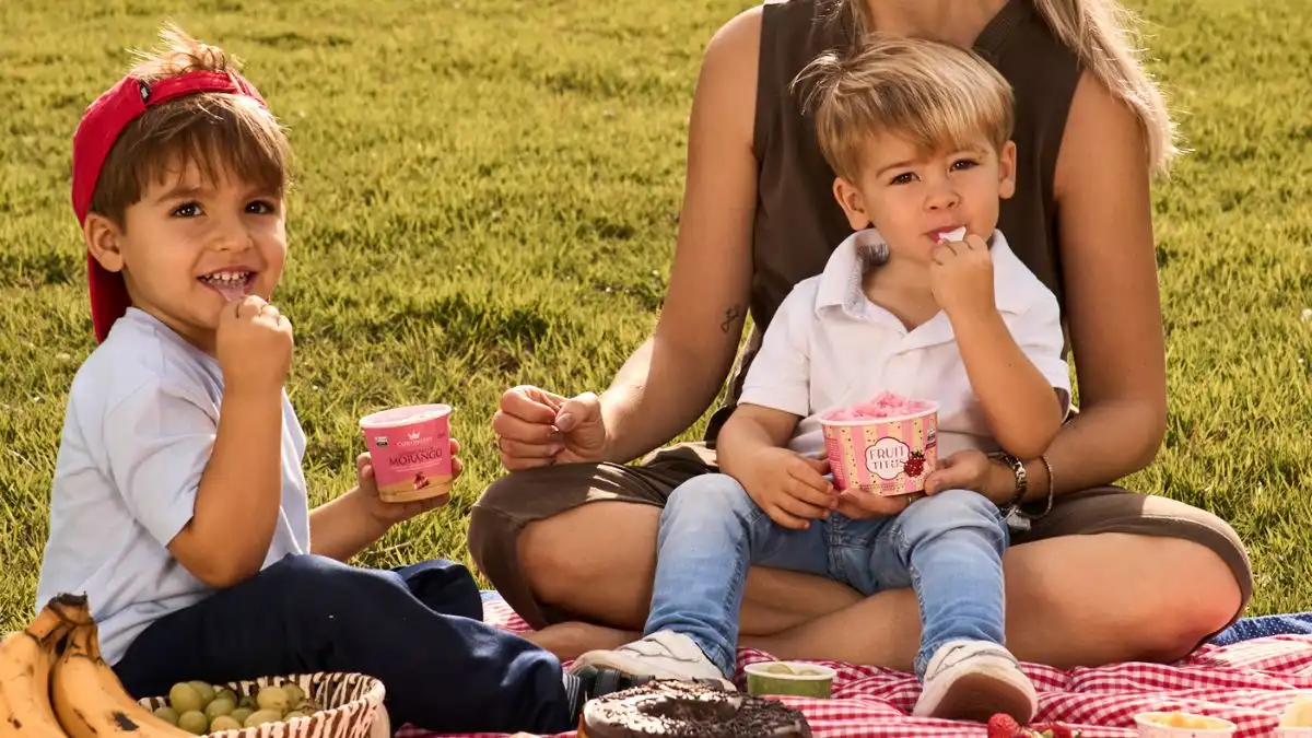 Crianças e uma mulher curtindo um piquenique ao ar livre, potes de 150ml com frutas, doces e uma atmosfera de alegria e diversão no parque.