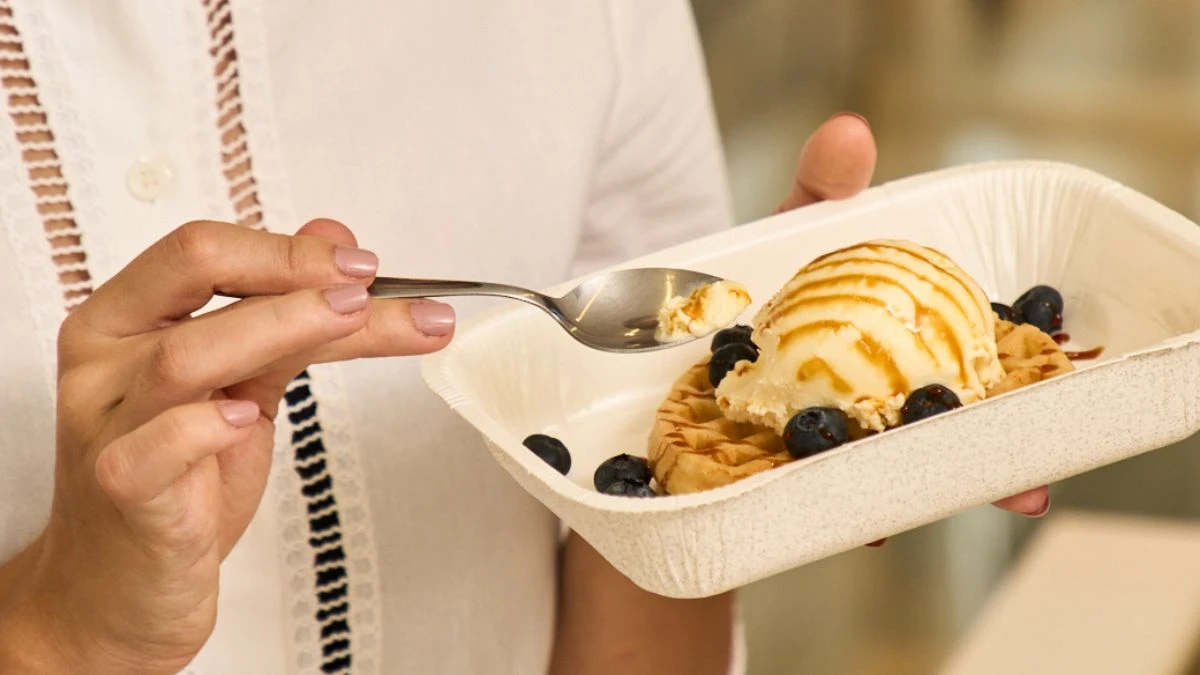 Pessoa segurando uma vasilha de sorvete com uma porção de waffle com sorvete de baunilha, calda de caramelo e blueberries, no estilo sobremesa.