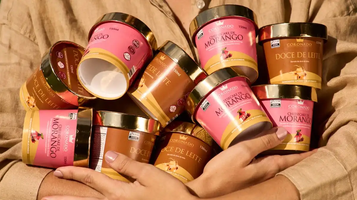 Variedade de pote de sorvete nos sabores de doce de leite, Morango e Morango com Chocolate. Acompanhados por mãos de pessoas ao fundo, ambientado em uma superfície de tecido.