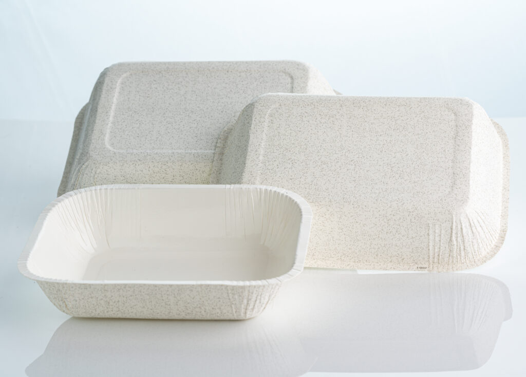 Três embalagens descartáveis ecológicas para delivery de alimentos. A imagem mostra uma bandeja retangular aberta em primeiro plano com interior branco liso e textura externa pontilhada, ao lado de outras duas embalagens viradas para baixo exibindo o fundo texturizado, sobre uma superfície clara e reflexiva com fundo branco.