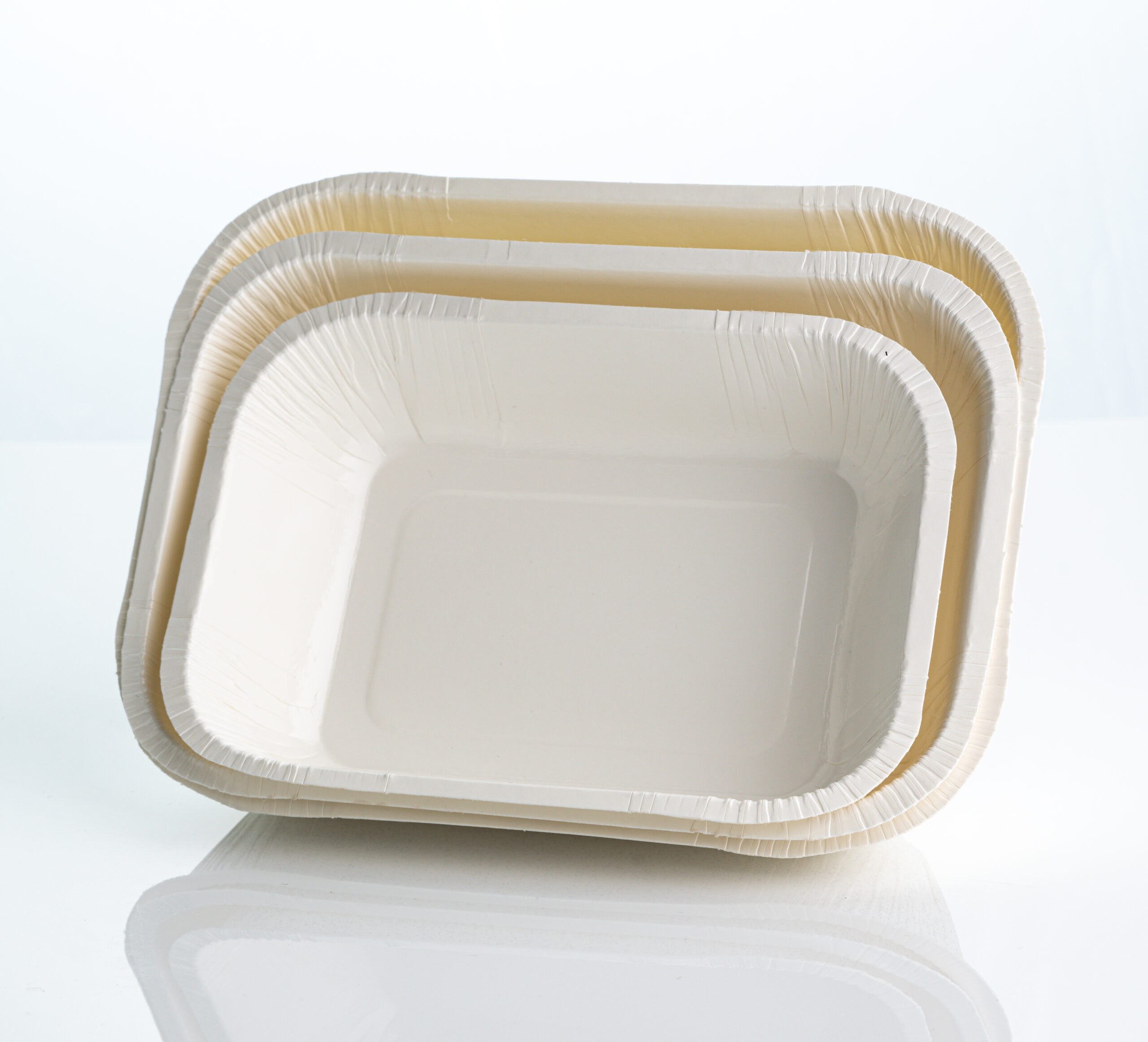 Três tamanhos de embalagens de papelão biodegradáveis e descartáveis para alimentos, no formato de bandejas retangulares brancas, empilhadas sobre uma superfície reflexiva clara com fundo branco.